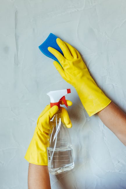 Close-up of a person wearing bright yellow rubber gloves cleaning a white wall with a blue sponge in one hand and a spray bottle of cleaning solution in the other, demonstrating surface cleaning and s